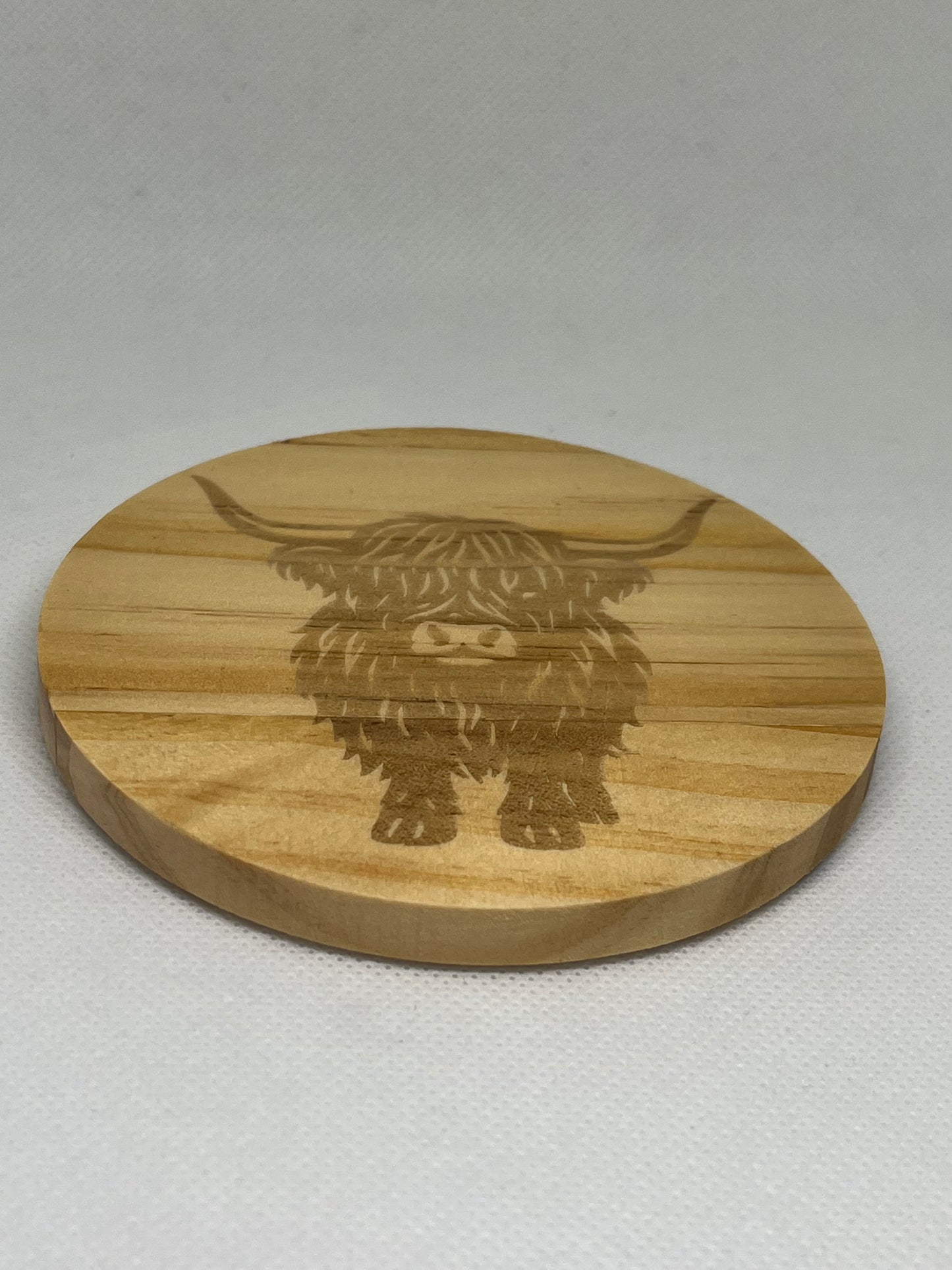 Round wooden coaster with a engraved highland cow design on a light gray background