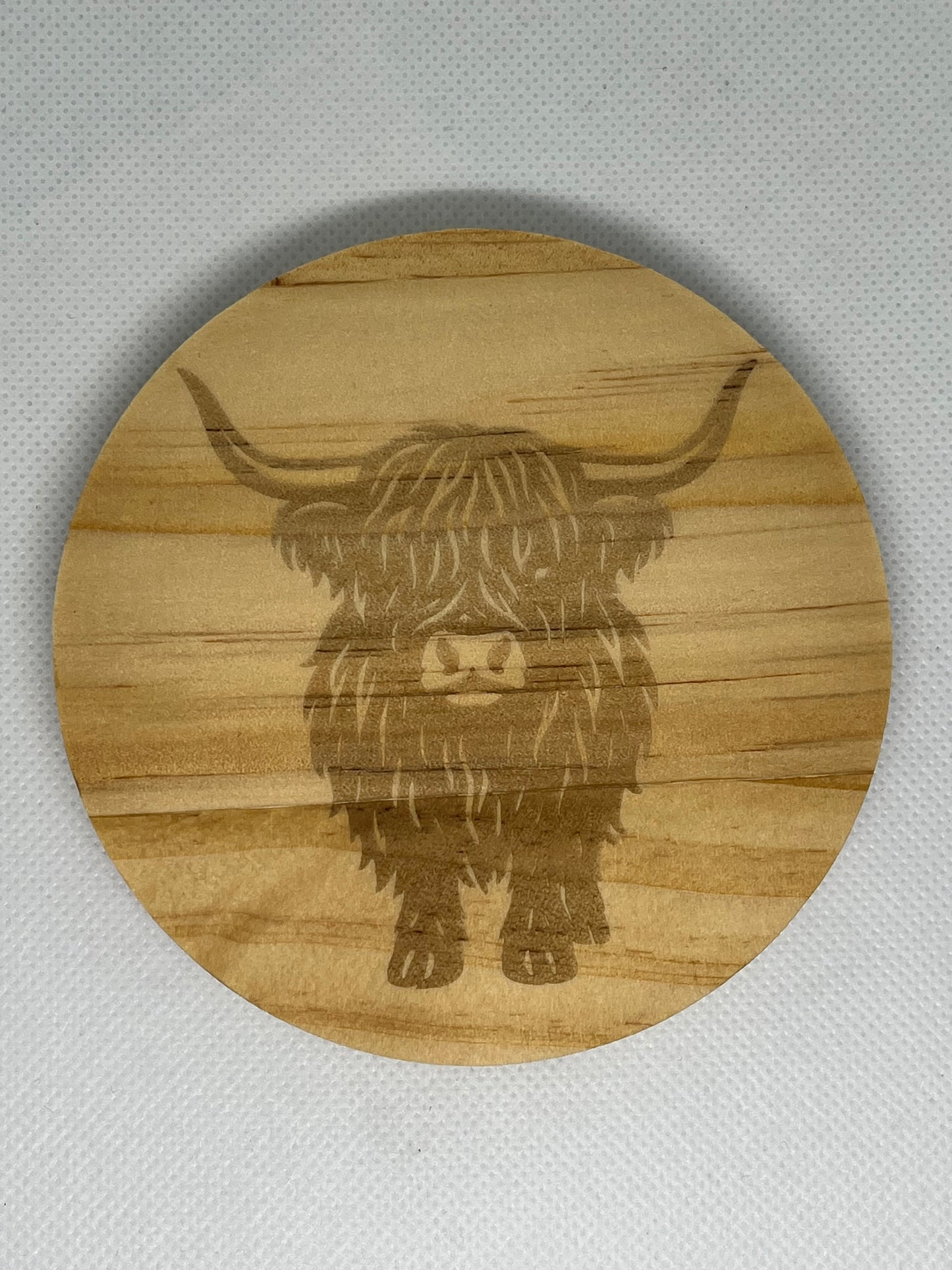 Round wooden coaster with engraved highland cow design on a gray background