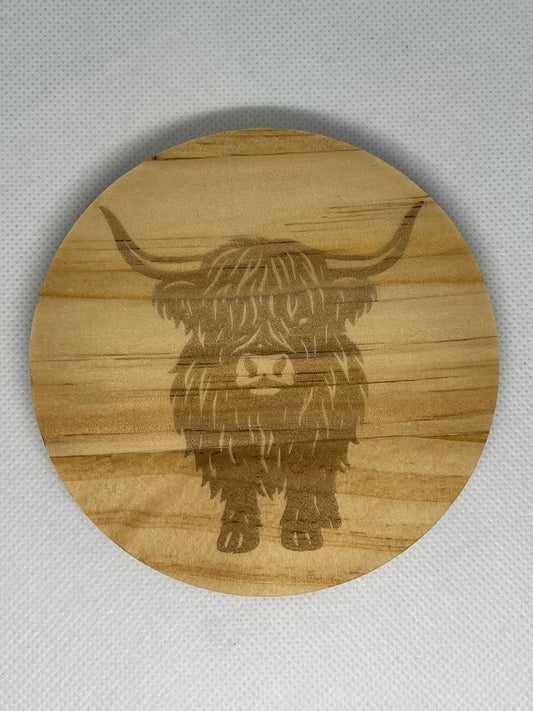 Round wooden coaster with engraved highland cow design on a gray background